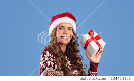Sexy young lady in Santa hat and warm sweater holding Christmas gift on blue studio background, panorama. Millennial woman with Xmas present for winter holidays posing and smiling 134414101