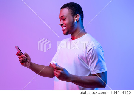 Tech Gadgets For Entertainment. Happy Black Guy Using Smartphone And Earpods For Listening Music, Holding Earphones Case In Hand, Cheerful African Man Standing In Neon Light Over Purple Background Tech Gadgets For Entertainment. Happy Black Guy Using Smartphone And Earpods For Listening Music, Holding Earphones Case In Hand, Cheerful African Man Standing In Neon Light Over Purple Background 134414140