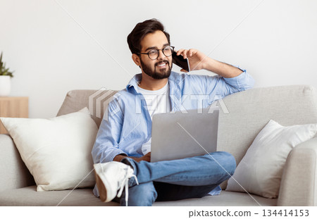 Remote Job Opportunities. Eastern Freelancer Man Talking On Cellphone And Using Laptop At Home, Smiling Arab Millennial Guy Working Online With Computer While Sitting On Couch In Living Room Remote Job Opportunities. Eastern Freelancer Man Talking On Cellphone And Using Laptop At Home, Smiling Arab Millennial Guy Working Online With Computer While Sitting On Couch In Living Room 134414153