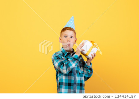 Birthday celebration, birthday gift concept. Excited ginger boy in festive paper cap holding gift box next to his ear, guessing whats inside, yellow studio background, copy space 134414187