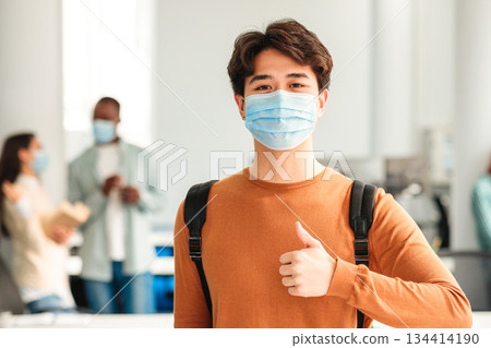 Protection Concept. Portrait of asian teenager wearing disposable surgical face mask to prevent infection, illness or covid-19, showing thumbs up sign gesture standing in classroom at high school Protection Concept. Portrait of asian teenager wearing disposable surgical face mask to prevent infection, illness or covid-19, showing thumbs up sign gesture standing in classroom at high school 134414190