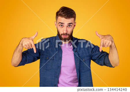 Surprised bearded guy pointing downwards on orange studio background, panorama. Emotional Caucasian guy showing or advertising something, offering big sale or product promotion 134414240