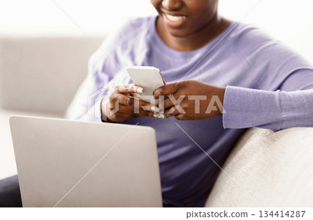 Scrolling News Feed. Closeup of unrecognizable african american smiling lady holding cellphone, typing sms message, sitting on couch with laptop. Black woman browsing internet, surfing web, using app 134414287