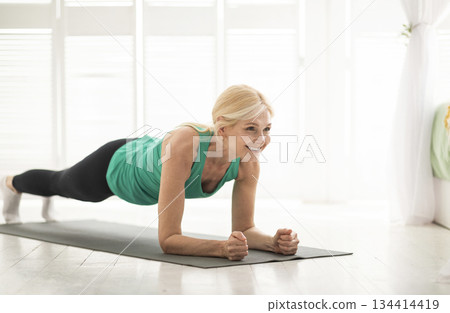 Home training concept. Smiling senior woman doing elbow plank on yoga mat in living room, sporty mature lady doing fitness workout routine, exercising indoors, strengthening her core muscles 134414419