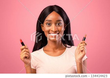 Portrait of happy black lady holding lipstick tubes over pink studio background. Charming African American woman choosing makeup, applying cosmetic product. Beauty and visage concept 134414423