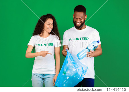 Don't Litter. Male And Female Multiethnic Volunteers Putting Used Plastic Bottle To Blue Garbage Bag Standing Over Green Studio Background. Keep Environment Clean, Ecology And Volunteering Work 134414430
