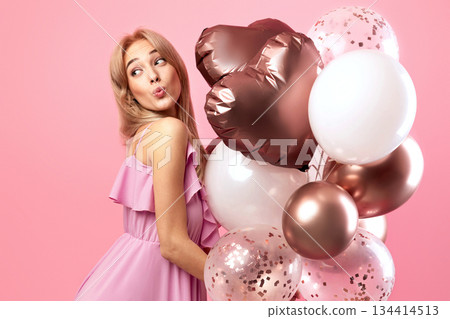 Beautiful blonde woman in cute dress holding balloons, pursing her lips for kiss, being playful on pink studio background, banner design with blank space. Party time, holiday celebration concept 134414513