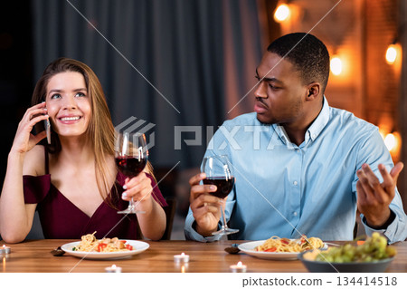 Unhappy Black Man Looking At Girlfriend Talking On Cellphone During Date In Restaurant, Feeling Angry And Annoyed. Young Multicultural Couple Having Communication Problems, Free Space 134414518