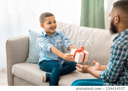 African Little Boy Giving Birthday Gift To Father Sitting Together On Sofa Celebrating Family Holiday At Home. B-Day Celebration. Gifts And Presents Concept. Selective Focus, Cropped African Little Boy Giving Birthday Gift To Father Sitting Together On Sofa Celebrating Family Holiday At Home. B-Day Celebration. Gifts And Presents Concept. Selective Focus, Cropped 134414537
