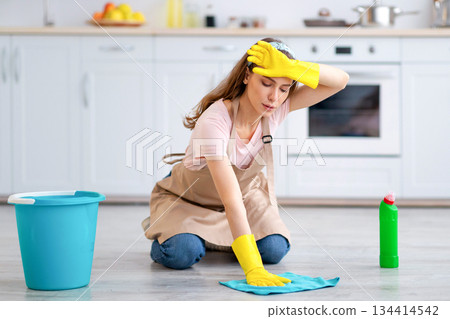 Full length portrait of tired millennial housewife washing floor, wiping her forehead at kitchen. Pretty young woman doing home cleanup, keeping her apartment tidy, feeling overworked 134414542