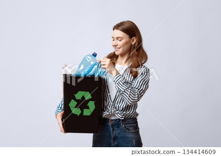 Waste Recycling Concept. Smiling Young Woman Holding Black Container With Plastic Bottles And Green Recycle Sign, Millennial Eco-Friendly Lady Standing Over Gray Background In Studio, Copy Space 134414609
