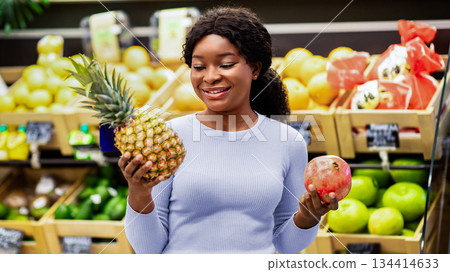 Portrait of positive black lady choosing fruits at grocery store or supermarket, holding pineapple and pomegranate. Young African American woman making selection of food at mall Portrait of positive black lady choosing fruits at grocery store or supermarket, holding pineapple and pomegranate. Young African American woman making selection of food at mall 134414633