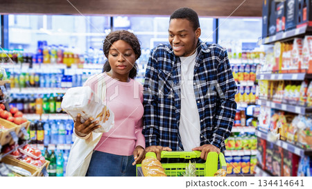 Portrait of happy black couple with shopping cart full of products choosing food together at modern supermarket. Millennial African American guy and his girlfriend buying groceries at store 134414641