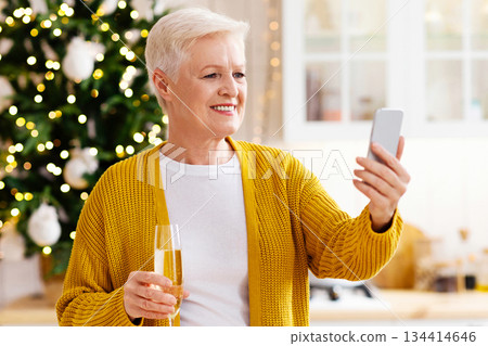 Beautiful grandmother having online party with family or friends on mobile phone during Christmas or New Year celebration, standing by xmas tree and drinking champagne, kitchen interior Beautiful grandmother having online party with family or friends on mobile phone during Christmas or New Year celebration, standing by xmas tree and drinking champagne, kitchen interior 134414646