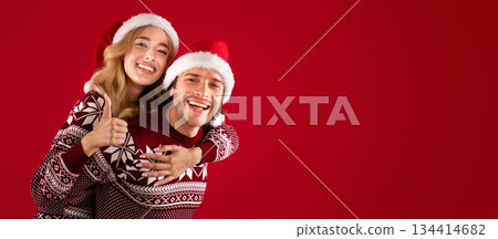 Xmas celebration. Millennial guy in Santa hat giving his girlfriend piggyback ride on red background Xmas celebration. Millennial guy in Santa hat giving his girlfriend piggyback ride on red background 134414682