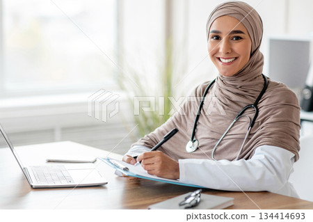 Smiling muslim woman doctor in headscarf taking notes in medical chart, sitting at workdesk with laptop at her cabinet in clinic, taking anamnesis while having conversation with patient, free space 134414693