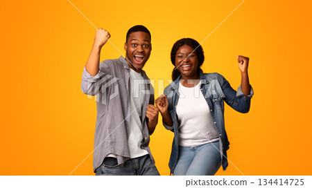 Lucky Winners. Overjoyed Young Black Man And Woman Raising Fists And Exclaiming With Excitement, Euphoric African American Couple Emotionally Celebrating Success Over Yellow Background, Free Space Lucky Winners. Overjoyed Young Black Man And Woman Raising Fists And Exclaiming With Excitement, Euphoric African American Couple Emotionally Celebrating Success Over Yellow Background, Free Space 134414725
