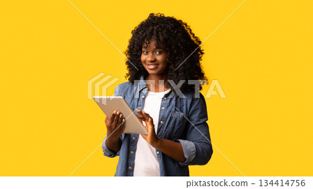 Cheerful beautiful black woman using modern digital tablet over yellow studio background, copy space. Cute african american young lady testing new mobile application on pad 134414756