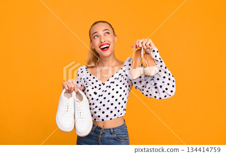 Studio portrait of a smiling stylish woman holding pair of high heels shoes and white sneakers in hands, choosing what to wear, looking up, isolated over pastel orange background wall, copy space 134414759