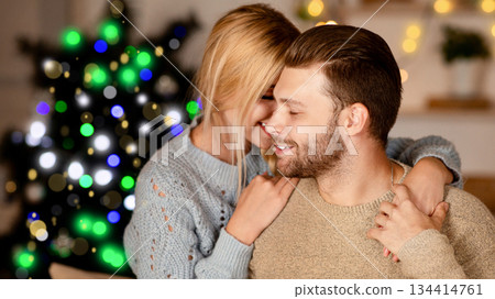 Young lovers cuddling at home, celebrating Christmas together, free space. Romantic couple having great time on xmas eve, sitting by Christmas tree, embracing and talking, love and affectionate 134414761