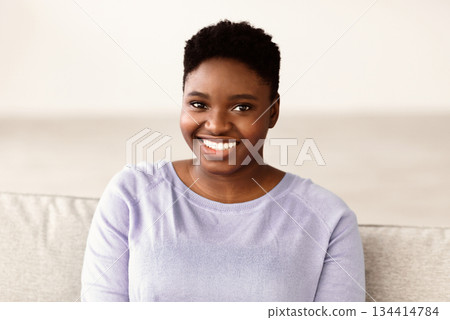 Natural Beauty. Headshot portrait of casual young large build black woman smiling, looking and posing at camera, resting alone on the sofa at home in bedroom. Closeup of positive chubby female Natural Beauty. Headshot portrait of casual young large build black woman smiling, looking and posing at camera, resting alone on the sofa at home in bedroom. Closeup of positive chubby female 134414784