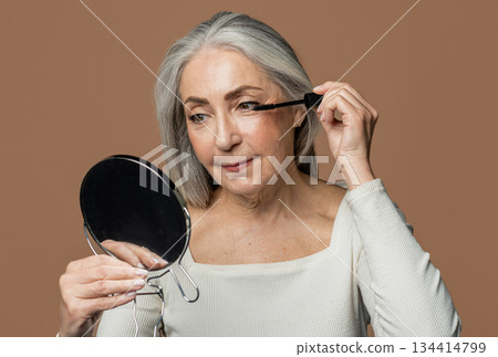 Cheerful senior caucasian female with gray hair applies mascara on eyelashes with brush, looks in mirror, isolated on brown background, studio. Lifestyle, beauty care, natural makeup, daily cosmetics 134414799