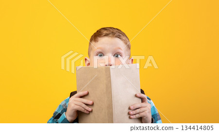 Shocked boy covering his mouth with book, reading exciting stories, yellow studio background, empty space. Emotional smart kid reading book, got impressed by novel. Educational concept Shocked boy covering his mouth with book, reading exciting stories, yellow studio background, empty space. Emotional smart kid reading book, got impressed by novel. Educational concept 134414805