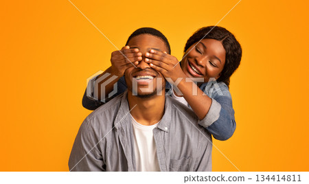 Happy african woman surprising her excited boyfriend, covering his eyes from the back, loving woman playing guess who with cheerful black man while they standing isolated over yellow background Happy african woman surprising her excited boyfriend, covering his eyes from the back, loving woman playing guess who with cheerful black man while they standing isolated over yellow background 134414811