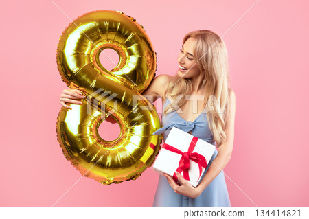 Happy young lady holding number eight golden balloon and bouquet of fresh spring tulips on pink studio background. 8th of March celebration, Woman's Day holiday concept Happy young lady holding number eight golden balloon and bouquet of fresh spring tulips on pink studio background. 8th of March celebration, Woman's Day holiday concept 134414821