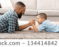 Raising A Real Man. Happy African Father And Son Arm Wrestling Competing Lying On Floor And Having Fun At Home. Little Boy Armwrestling With Daddy. Family Bond Concept. Side View 134414842
