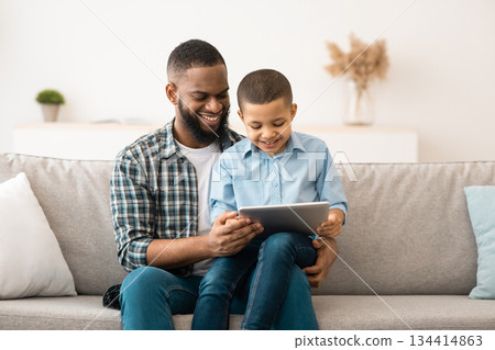 African American Father And Son Using Tablet Computer Browsing Internet Together Sitting On Couch At Home. Weekend Family Leisure, Gadgets And Modern Lifestyle Concept 134414863