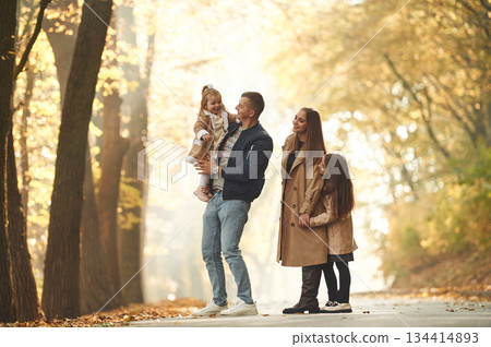 Holding girl in hands. Happy family of mother, father and two daughters are in the autumn park, forest, road 134414893