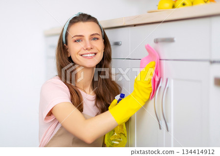 Positive young lady washing kitchen cabinet with detergent and rag, smiling at camera indoors. Lovely millennial housemaid doing chores, keeping her home tidy, performing cleanup Positive young lady washing kitchen cabinet with detergent and rag, smiling at camera indoors. Lovely millennial housemaid doing chores, keeping her home tidy, performing cleanup 134414912