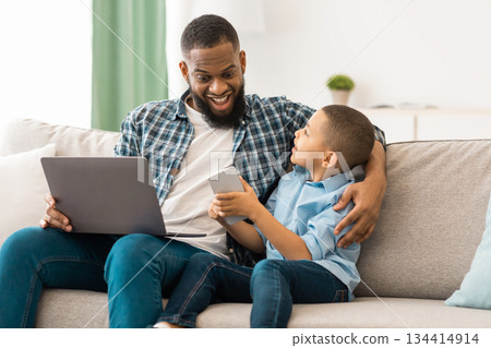 Black Father And Son Using Gadgets, Boy Showing Smartphone To Excited Dad Sitting With Tablet Computer And Browsing Internet Together At Home. Technology, Devices And Modern Family Lifestyle 134414914