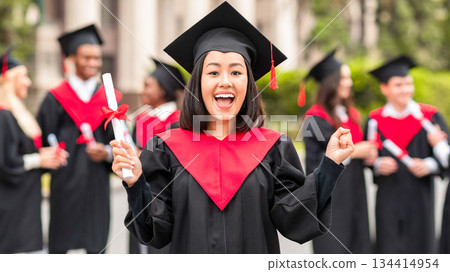 Delighted asian young lady in graduation costume showing her diploma and screaming at camera, happy female student posing over international group of students at university campus, copy space Delighted asian young lady in graduation costume showing her diploma and screaming at camera, happy female student posing over international group of students at university campus, copy space 134414954