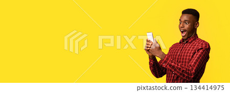 Mobile Offer. Surprised funny black guy looking at smartphone screen with excitement, emotionally reacting to online news, standing with opened mouth isolated over yellow background, copy space 134414975