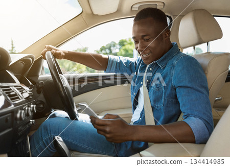 Smiling black guy using smartphone, answering messages or checking emails while driving car, side view, copy space, sun flare. Afro american driver checking his cellphone, dont look at road Smiling black guy using smartphone, answering messages or checking emails while driving car, side view, copy space, sun flare. Afro american driver checking his cellphone, dont look at road 134414985