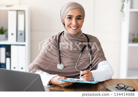 Smiling young woman doctor in hijab taking anamnesis in clinic, sitting in front of laptop, holding medical chart and pen, copy space. Female therapist in head scarf smiling at camera while working 134414986