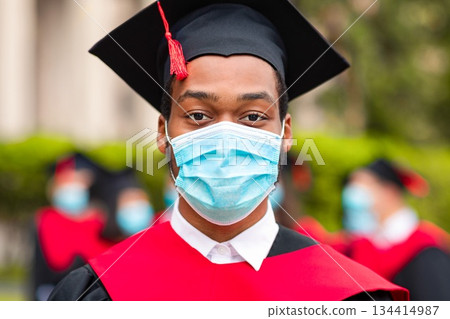 African american guy in graduation costume and protective face mask, black man student posing over international group of students at university campus, 2021 graduation concept 134414987