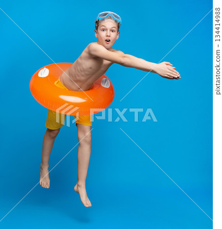 Teen boy in inflatable ring about to dive into swimming pool, blue studio background Teen boy in inflatable ring about to dive into swimming pool, blue studio background 134414988