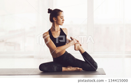 A young woman is depicted sitting on a yoga mat in a well-lit studio. She is dressed in black athletic wear and is performing a yoga pose, maintaining a focused yet serene expression 134415000