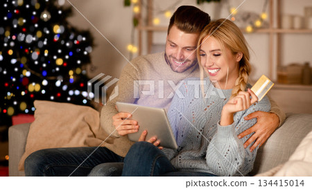 Delighted husband and wife making purchases online, using digital tablet and credit card, Christmas atmosphere at home. Happy young family shopping online on pad, using Christmas offers, copy space 134415014