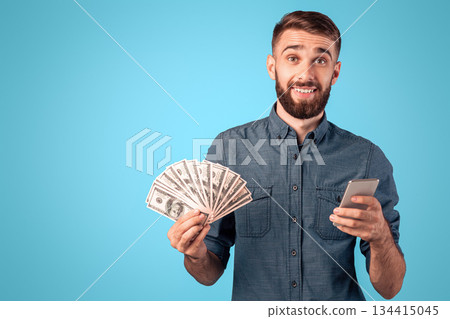 Online bank credit, lottery, casino. Happy young guy holding heap of money and smartphone on blue studio background. Rich millennial man with dollar banknotes and cellphone achieving financial success Online bank credit, lottery, casino. Happy young guy holding heap of money and smartphone on blue studio background. Rich millennial man with dollar banknotes and cellphone achieving financial success 134415045