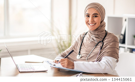 Smiling muslim woman doctor in headscarf taking notes in medical chart, sitting at workdesk with laptop at her cabinet in clinic, taking anamnesis while having conversation with patient, free space Smiling muslim woman doctor in headscarf taking notes in medical chart, sitting at workdesk with laptop at her cabinet in clinic, taking anamnesis while having conversation with patient, free space 134415061