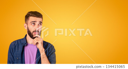 Portrait of pensive young bearded man touching his chin, deep in thought over orange studio background. Doubtful millennial guy thinking and making decision, dreaming about something 134415065