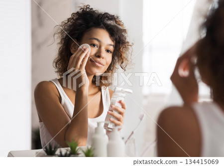 Young afro-american woman cleaning face with cotton pad looking in mirror at bathroom 134415069