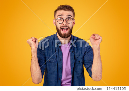 Winner and success concept. Excited young guy making YES gesture with clenched fists on orange studio background. Excited Caucasian man shouting OMG, overjoyed with big win 134415105