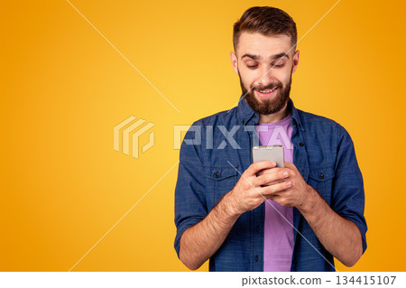 Handsome bearded guy with smartphone browsing internet, communicating online or shopping over orange background. Young man working or studying remotely using mobile device Handsome bearded guy with smartphone browsing internet, communicating online or shopping over orange background. Young man working or studying remotely using mobile device 134415107