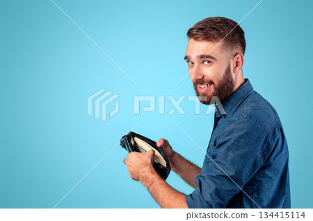 Funky Caucasian guy holding full wallet with money on blue studio background. Wealthy young man with purse of dollars, achieving financial success, receiving raise of salary or profit 134415114