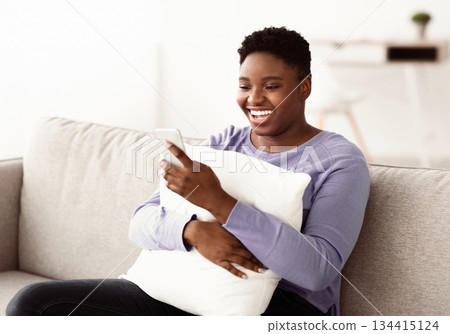 Domestic Rest And Free Time. Body positive black woman using smartphone, making video call, holding and hugging pillow, sitting on couch in living room. Lady browsing internet, relaxing at home Domestic Rest And Free Time. Body positive black woman using smartphone, making video call, holding and hugging pillow, sitting on couch in living room. Lady browsing internet, relaxing at home 134415124